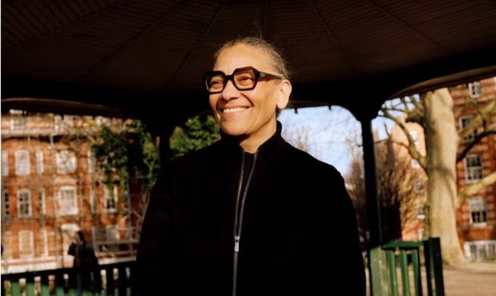a black woman wearing a black top and glasses smiles whilst standing in a bandstand with trees behind her